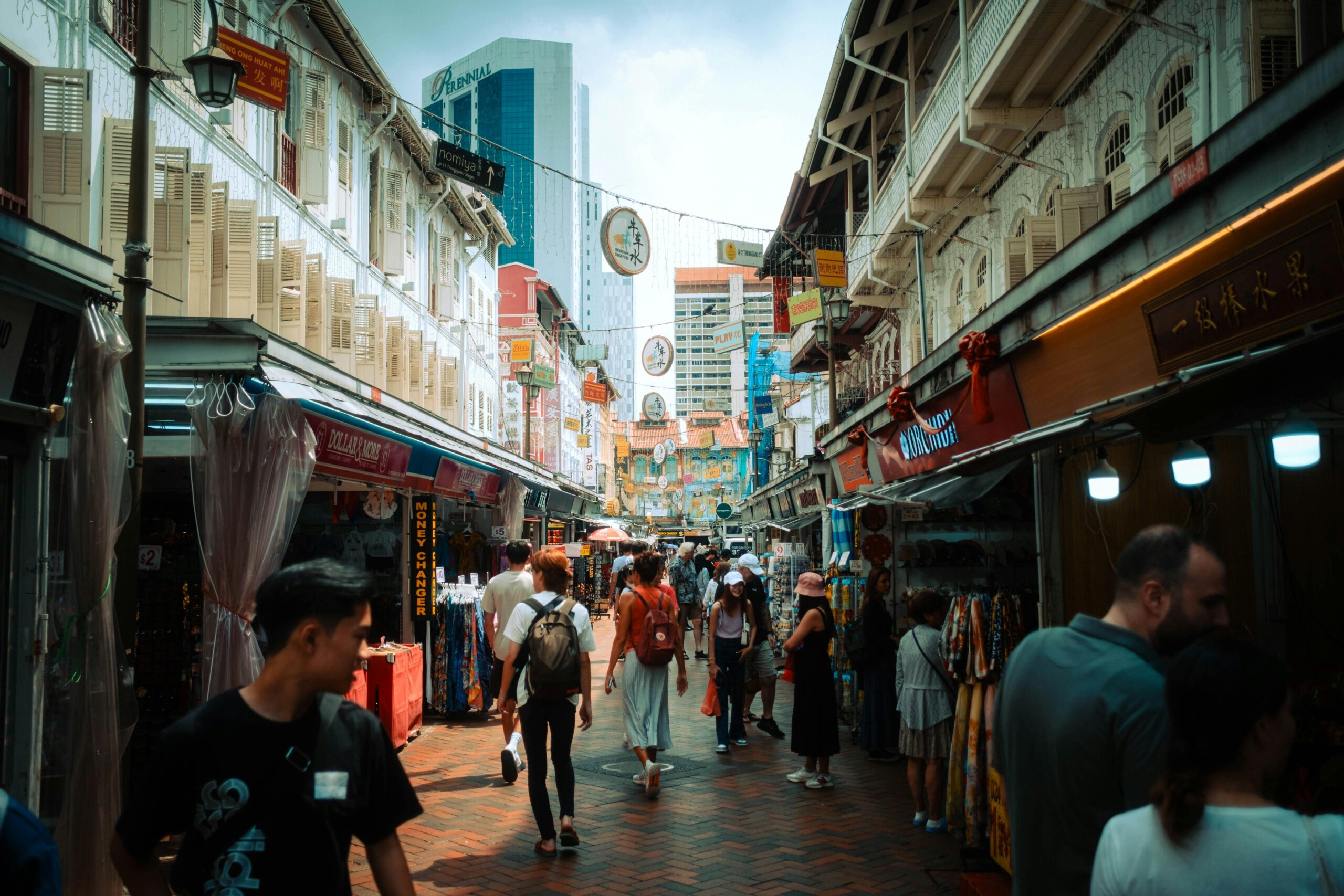 A crowded, colorful marketplace flanked by buildings with traditional architecture, with a modern skyscraper visible in the background under a partly cloudy sky.