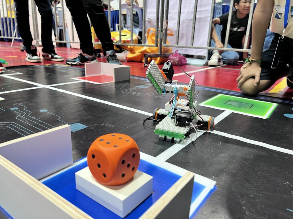 At Camp Cosmos, an educational robotic vehicle maneuvers expertly on a grid, skillfully navigating between obstacles with a large orange die nearby, while curious summer camp participants observe in the background.