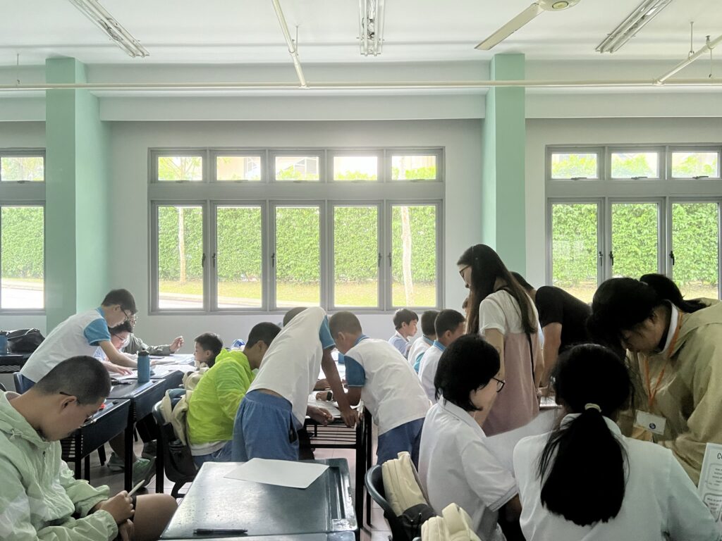 A classroom at Education Summer Camp in Singapore is bustling with students in uniforms working at desks, some standing and some seated. A teacher from Camp Cosmos assists a group. Large windows line one wall, illuminating the energetic scene.