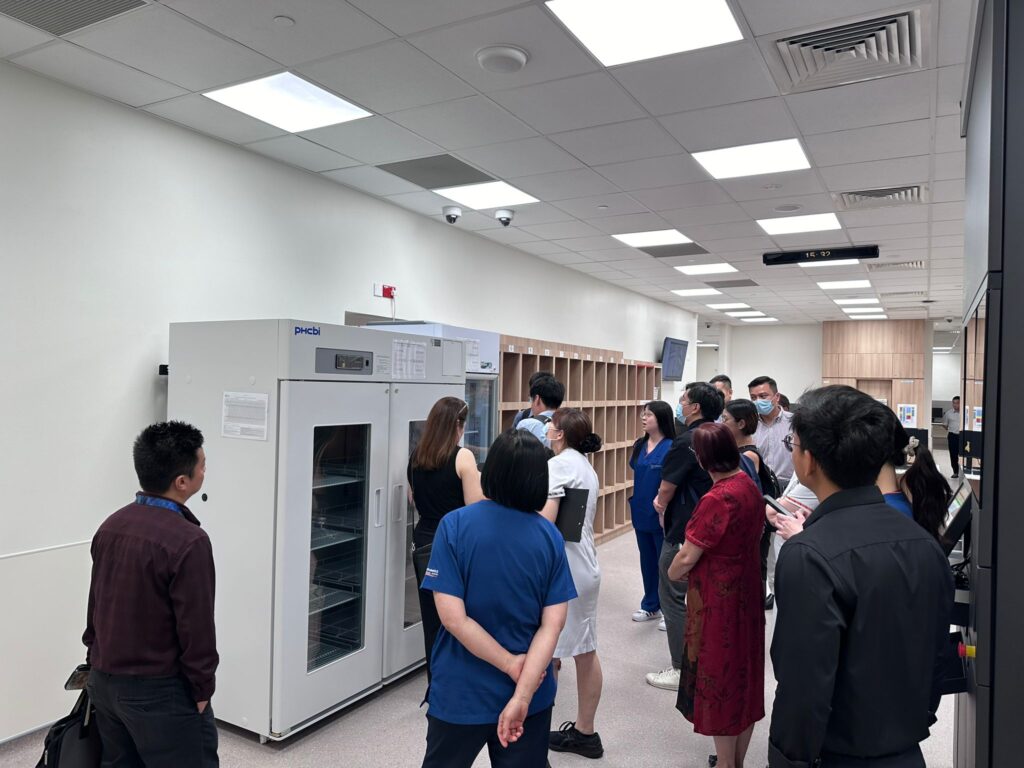 A group of people stand in a room, observing a large piece of equipment in a lab or medical setting.