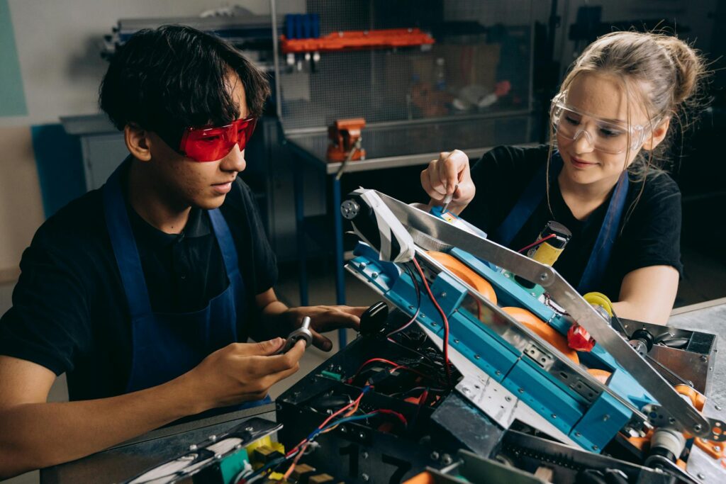 At Camp Cosmos, two people wearing safety goggles work on a blue and orange robotic machine at a table, surrounded by tools and wires. This educational experience combines hands-on learning with Summer Camp fun.